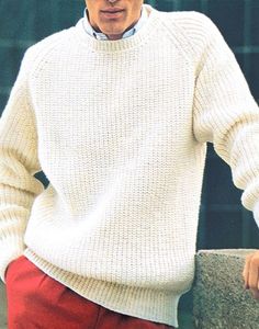 a man in a white sweater and red pants leaning against a stone wall with his hands on his hips