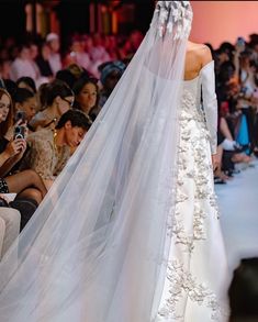 the back of a wedding dress with a veil on it's head and people in the background