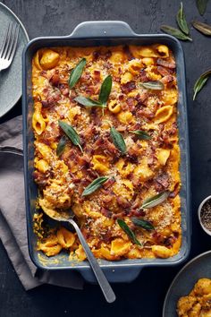 a casserole dish with pasta, bacon and sage leaves in it on a table