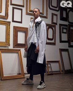 a man in white shirt and black skirt standing next to framed pictures on wall with gold frames