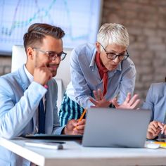 three people in business attire look at a laptop screen while sitting at a table with pens and pencils