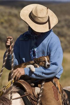 a man riding on the back of a horse with a dog in his lap and wearing a cowboy hat