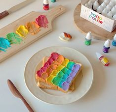 a rainbow cake on a white plate next to some paint and wooden utensils