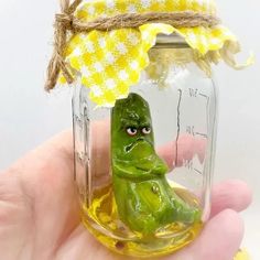 a hand holding a jar filled with yellow liquid and a green frog figurine