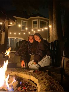 two women sitting next to each other in front of a fire pit with lights on