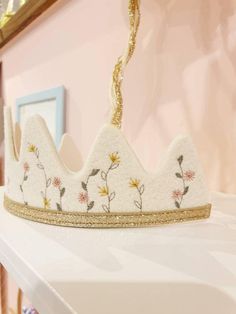 a white crown with flowers on it is hanging from a gold chain in front of a pink wall