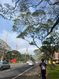 a person walking down the street with cars parked on both sides and a rainbow in the sky