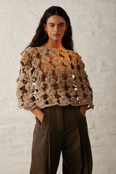 a woman standing in front of a white wall wearing brown pants and a crocheted top