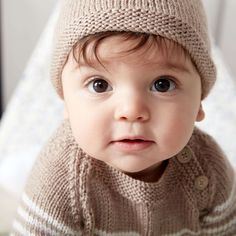 a baby wearing a knitted sweater and beanie looks at the camera while sitting on a bed