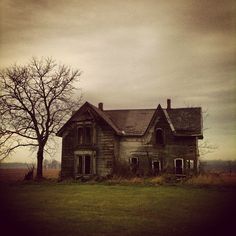 an old house sitting in the middle of a field with a tree next to it