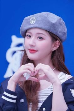 the girl is wearing a hat and making a heart shape with her hands while standing in front of a blue wall