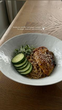 a white bowl filled with noodles and meat next to cucumbers on top of a wooden table