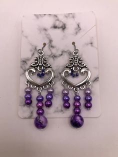 2 Inch drop-earrings made with hypoallergenic materials.   Beads are attached to a silver plated heart charm consisting of a purple marble bead and three smaller glass beads in three different shades of purple/lavender in the center.  Left/Right sides contain the same smaller glass beads in the three different shades of purple/lavender as the center.  The middle of the heart charm is accented with four tiny glass beads as an accent.  These beads are a darker shade of purple. Includes soft ear backings. Materials are environmental, lead free and non-toxic. Light weight, smooth surface, and a touch of sheen to add elegance. The earrings are attached to a marble cardboard earring holder and are shipped in a sealed plastic cover, and placed inside a mesh drawstring "thank you" pouch as shown. Purple Metal Dangle Earrings, Purple Dangle Metal Earrings, Purple Dangle Heart Earrings For Pierced Ears, Purple Teardrop Metal Earrings, Nickel-free Purple Chandelier Earrings Gift, Dangle Heart Bead Earrings, Metal Dangle Heart Earrings With Heart Beads, Purple Metal Earrings, Metal Heart Dangle Earrings With Heart Beads