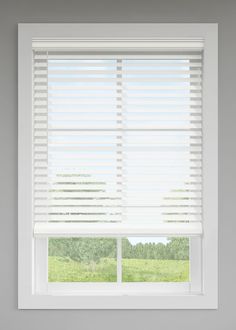 a window with white blinds in front of a green grass covered field and trees behind it