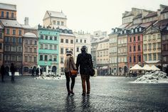 two people walking down a cobblestone street in the snow with buildings behind them