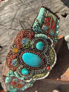 This beautiful bead embroidered cuff features a large genuine turquoise cabochon at the center, flanked by 2 smaller turquoise cabochons on either side.  The beadwork surrounding the cabochons is worked in a style that emulates the beautiful floral beadwork of the Ojibwe tribes of the Pacific Northwest.  All beads are hand stitched (2 beads at a time) into a felt foundation, and backed by ultrasuede.  Flanking each side of the cuff are 2 pieces of loomed beadwork in a pattern that I designed to compliment the center beadwork.  These are mounted on a beautiful sturdy piece of 2-tone taffeta that is beaded with aged turquoise glass beads from Portugal.  The closure of this cuff incorporates a lovely turquoise colored glass button from the Czech Republic and a deerskin thong that loops around Handmade Turquoise Artisan Cuff Bracelet, Artisan Turquoise Beaded Bracelet, Handmade Artisan Turquoise Cuff Bracelet, Bohemian Turquoise Bracelet With Gemstone Accents, Bohemian Turquoise Bracelets With Gemstone Accents, Handmade Artisan Turquoise Beads, Artisan Beaded Turquoise Cuff Bracelet, Artisan Turquoise Beaded Cuff Bracelet, Elegant Turquoise Beaded Gems And Cabochons