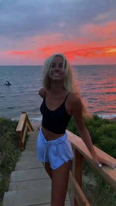 a woman standing on a wooden railing next to the ocean at sunset with her arms behind her back