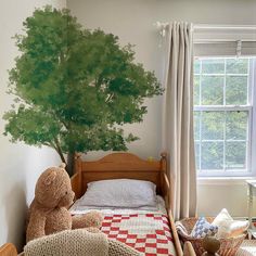 a bedroom with a tree painted on the wall and teddy bear sitting in bed next to it