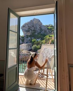 a woman sitting in front of an open window with a painting on the easel