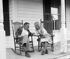 an old man and woman sitting on rocking chairs talking to each other in front of a house