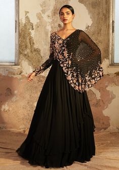 Editor's Note Black Double Layered Cape Style Georgette Gown With One Full Sleeve Featuring Gold 3d Floral Hand Embellishment. Color: Black Fabric: Georgette, Crepe Care: Spot Dry Clean Only About the Designer Shloka Khialani is a contemporary Indianwear label that entails a clean yet effective approach to design. Emphasizing on modern cuts and unconventional embellishments, the label aims to bring it’s minimal yet modern design philosophy to the forefront. Flowy silhouettes, an array of solid h Black Gown Indian, Hand Embellishment, Haldi Outfit, Georgette Gown, Cape Gown, Indo Western Dress, Cape Style, Black Layers, Desi Clothes