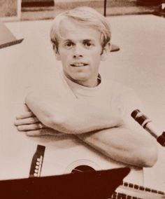 an old black and white photo of a young man with his arms crossed, leaning on a guitar