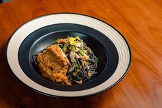 a black plate topped with pasta and meat on top of a wooden table next to a fork