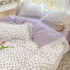 an unmade bed with purple and white comforter on it, next to a window