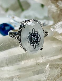 This filigree ring is an Edwardian antique reproduction in sterling silver set with a camphor glass with a sterling silver filigree center embellishment inset with your choice of white CZ, Moissanite, or genuine diamond. The inset center gem is 1mm to 1.5mm in diameter. This faceted oval gem is 14mm x 10mm. This ring sits 7mm off the finger. The inside of the band is marked 925 for solid sterling silver. Silver Moonstone Ring With Rose Cut Diamonds For Wedding, Anniversary White Gold Moonstone Ring With Diamond Accents, Silver Moonstone Ring With Center Stone For Wedding, Intricate Design Moonstone Jewelry, Silver Cubic Zirconia Moonstone Wedding Ring, Heirloom Silver Moonstone Jewelry, Spiritual Gemstones For Wedding, Heirloom Style Silver Ring With Moonstone, Vintage Oval Gemstones For Wedding