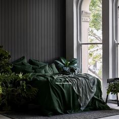 a bed with green sheets and pillows in front of an open window next to potted plants