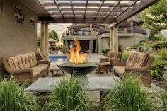 a fire pit sitting in the middle of a patio surrounded by grass and bushes with chairs around it