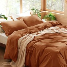 an unmade bed with brown sheets and pillows in front of a large window filled with potted plants