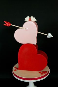 a red heart shaped cake with an arrow sticking out of it's center and two hearts on top