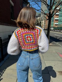 a woman standing on the sidewalk with her back to the camera, wearing jeans and a crocheted sweater