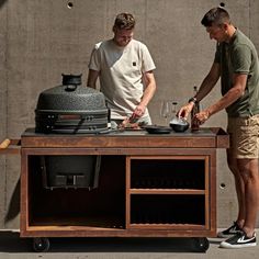 two men preparing food on top of a table with an outdoor grill in the background