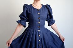 Vintage blue linen Austrian Folk dress with puffy sleeves from the 1990's. Women's traditional Oktoberfest dress made of navy blue linen fabric. The dress has a button down front with gold metal engraved and horn buttons. The bodice is fitted, has short volumetric sleeves with elastic hems. The skirt is pleated at the waist, has one hand pocket and falls free below the knees. Handmade in the 90s. No material or size indicating tags. Fabric feels like linen/cotton blend.  Size: Fits like 38/40 DE, which is 8/10 US or M/L. Model shown on the photos wears 38 DE / 8 US / M, bust size Cup A. To be sure this item would fit you please check the measurements below. Approximate Measurements (taken while dress is lying flat, double armpit to armpit and waist): Shoulders: 13.5 inches / 34.3 cm Sleeve Puffy Sleeve Dress, Oktoberfest Dress, Dress With Puffy Sleeves, Traditional Cottage, Dress Traditional, Folk Dress, Navy Blue Linen, Folk Dresses, Puffy Sleeves