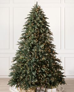 a small christmas tree with lights and presents under it on a wooden floor in front of a white wall