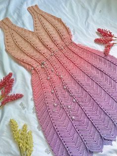 a pink and purple knitted dress sitting on top of a white blanket next to flowers