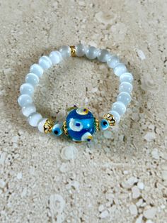 Evil Eye Bracelet featuring a Venetian Glass focal bead measuring approximately 20MM in shades of blue, white and yellow, accented with 6MM evil eye beads, gold plated bead caps and 8MM smooth round cat's eye beads.  Bracelet is strung on stretch cord and measures approximately 7 1/2 inches.  Handmade. GIFT BOXED White Spiritual Bracelets With Colorful Beads, White Round Bohemian Stretch Bracelet, Spiritual White Bracelets With Colorful Beads, White Bohemian Stretch Bracelet, White Glass Beaded Bracelets With Round Beads, White Glass Beaded Bracelets, White Beaded Round Evil Eye Bracelet, White Round Beaded Evil Eye Bracelet, White Evil Eye Bracelet With Round Beads