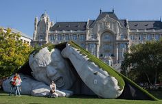 two people are standing in front of a giant sculpture with grass growing on it's head