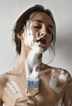 a woman with white paint on her face and body is holding a paintbrush in front of her face