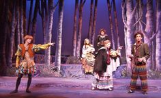 several people dressed in costumes on stage with trees and bushes behind them, one person pointing at something