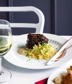 a white plate topped with meat and rice next to a glass of wine on a table