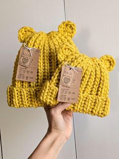 a hand holding two knitted hats with tags attached to them, one is yellow and the other is brown