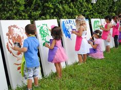 several children are painting on the side of white boards in front of a hedge wall