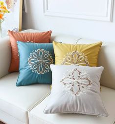 four different colored pillows on a white couch in front of a vase with yellow and orange flowers