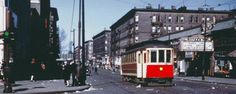 a red and white trolley car on street next to tall buildings with people standing around