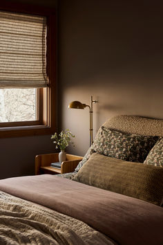 a bed sitting under a window next to a wooden table with a lamp on it