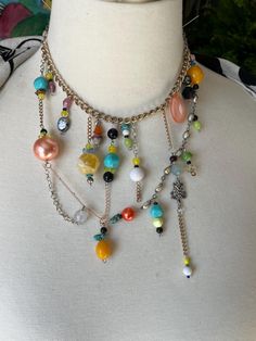 This one-of-a-kind necklace is a must-have for every fashion-forward woman. Handmade with a unique design, it features a vibrant and colorful mix of materials - turquoise, tigers eye, glass, plastic, paper, metal, and alloy. The statement piece is perfect for adding a pop of color to any outfit and is sure to turn heads with its head-turning style. The necklace is multi-chain and features 15 colorful charms, giving it a multi-fashion look. The mixed metallic chains, including faux rose gold, gold, and silver, add a touch of glamour to the piece. The lobster claw closure and adjustable length ensure a perfect fit for any wearer. This vogue 2025 style necklace is a true standout, making it a perfect gift for the fashionista in your life. Whether for a special occasion or everyday wear, it is Trendy Multi-strand Metal Charm Necklaces, Unique Multi-strand Colorful Beaded Necklaces, Multicolor Beaded Multi-strand Layered Necklace, Whimsical Multicolor Pendant Necklace, Whimsical Multicolor Necklaces With Charms, Dangly Necklace, Victorian Necklace, Eye Glass, Tigers Eye