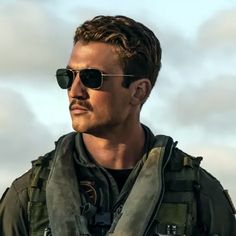 a man wearing sunglasses and a green jacket looks off into the distance with clouds in the background